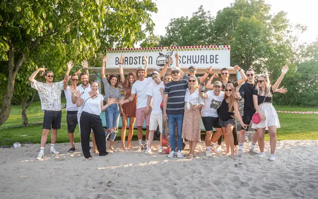 Team und Gäste jubeln vor dem Bordsteinschwalbe-Schild am Strand – Imagefilm-Dreh in Neuss